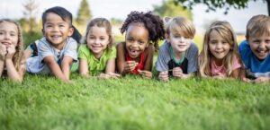 A large group of school aged children lay together in the grass on a sunny summer day as they pose for a portrait. They are each dressed casually and are smiling.